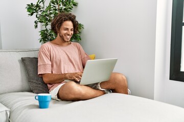 Young hispanic man smiling happy using laptop sitting on the sofa at home.