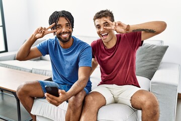 Young hispanic men using smartphone sitting on the sofa at home doing peace symbol with fingers over face, smiling cheerful showing victory