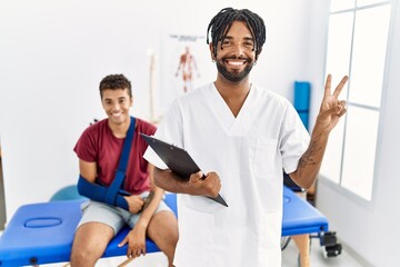 Young hispanic man working at pain recovery clinic with a man with broken arm smiling with happy face winking at the camera doing victory sign with fingers. number two.