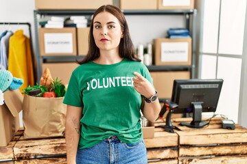 Young brunette woman wearing volunteer t shirt at donations stand pointing with hand finger to the side showing advertisement, serious and calm face