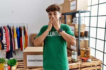 Young arab man wearing volunteer t shirt at donations stand laughing and embarrassed giggle covering mouth with hands, gossip and scandal concept