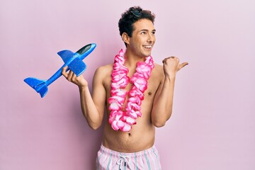 Young handsome man wearing swimsuit and hawaiian lei holding airplane toy pointing thumb up to the...