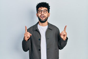 Young arab man with beard wearing glasses amazed and surprised looking up and pointing with fingers and raised arms.