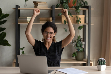 Euphoric mood. Excited young latin lady worker sit by desk raise hands up after successful finish...