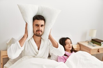 Man covering ears with pillow irritated for snore lying in bed at home.
