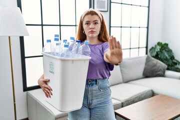 Young redhead woman holding recycling wastebasket with plastic bottles doing stop sing with palm of the hand. warning expression with negative and serious gesture on the face.