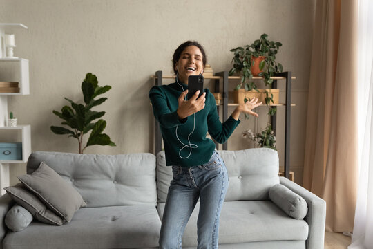 Full of joy. Young latin woman laughing kidding by music in earphones shooting selfie on cell in domestic atmosphere. Active positive millennial lady make video call pose for funny clip at living room