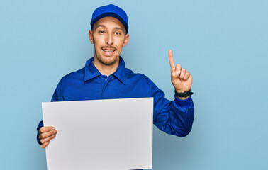 Bald man with beard wearing builder jumpsuit uniform holding empty banner surprised with an idea or question pointing finger with happy face, number one