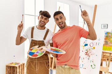 Two hispanic men couple smiling confident holding paintbrushes and palette at art studio