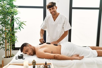 Two hispanic men physiotherapist and patient having back treatment using hot stones at beauty center