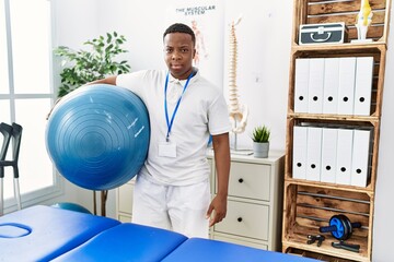 Fototapeta premium Young african physiotherapy man holding pilates ball at rehabilitation clinic thinking attitude and sober expression looking self confident