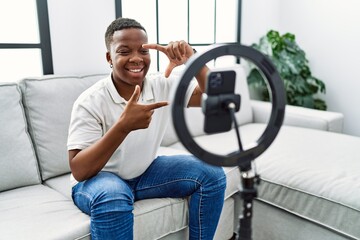 Young african man recording vlog tutorial with smartphone at home smiling making frame with hands and fingers with happy face. creativity and photography concept.