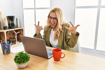Beautiful blonde woman working at the office with laptop shouting with crazy expression doing rock...