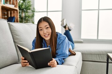 Young chinese girl reading book lying on the sofa at home.
