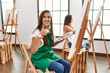 Young hispanic artist women painting on canvas at art studio smiling positive doing ok sign with hand and fingers. successful expression.