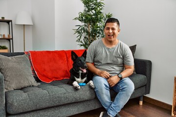 Young latin man and dog sitting on the sofa at home winking looking at the camera with sexy...