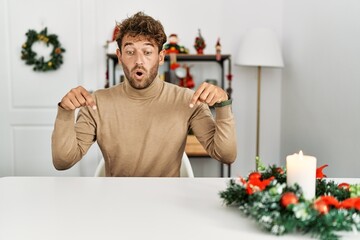 Young handsome man with beard sitting on the table by christmas decoration pointing down with fingers showing advertisement, surprised face and open mouth