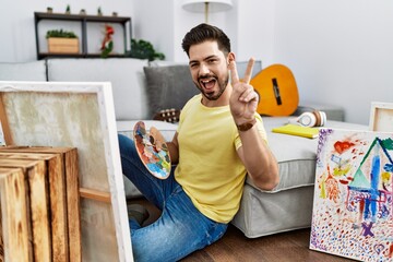 Young man with beard painting canvas at home smiling with happy face winking at the camera doing...