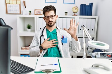 Young man with beard wearing doctor uniform and stethoscope at the clinic swearing with hand on...