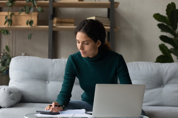 Doing calculations. Confident millennial latin female housewife focused on paperwork use calculator to check online utility bills payment. Young woman control expenditures count total cost by receipts