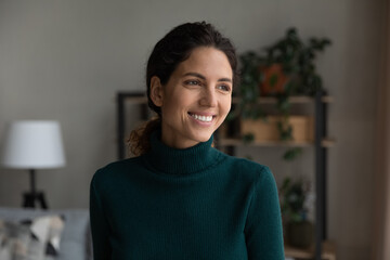 Beautiful smile. Head shot of smiling carefree hispanic female standing at living room looking aside lost in positive optimistic thoughts. Happy young lady enjoying life dreaming visualizing at home