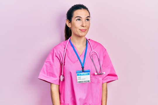 Young brunette woman wearing doctor uniform and stethoscope smiling looking to the side and staring away thinking.