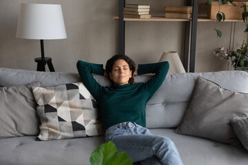 Relaxing at cozy home. Serene casual latina female recline with hands over head on comfy sofa at...