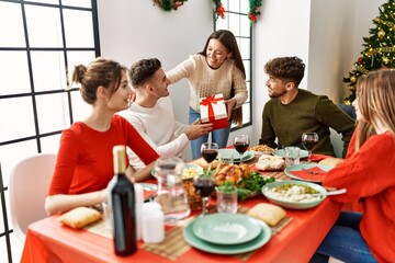 Group of young people smiling happy having christmas dinner. Woman giving gift to man at home.