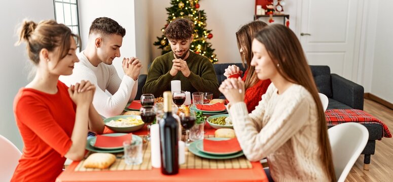 Group of young people celebrating christmas praying for food sitting on the table at home.