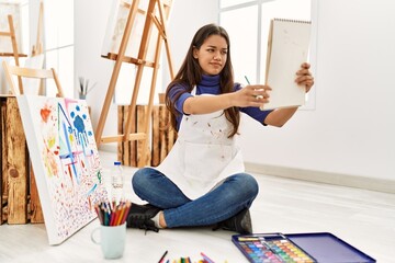 Young latin woman with doubt expression looking notebook draw at art studio
