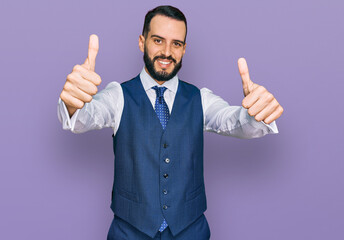 Young man with beard wearing business vest approving doing positive gesture with hand, thumbs up smiling and happy for success. winner gesture.