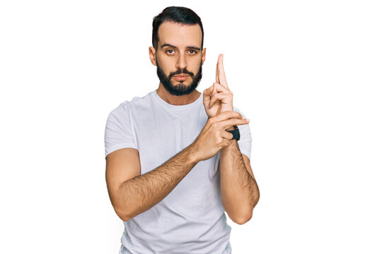 Young man with beard wearing casual white t shirt holding symbolic gun with hand gesture, playing killing shooting weapons, angry face