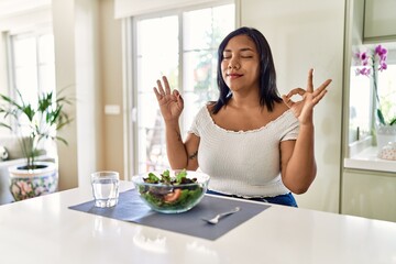 Young hispanic woman eating healthy salad at home relax and smiling with eyes closed doing meditation gesture with fingers. yoga concept.