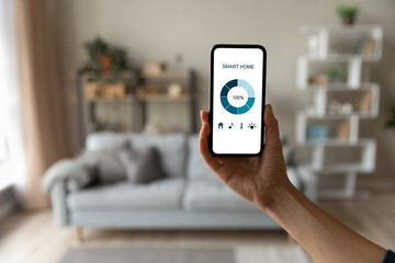 Internet of things. Focus on modern smartphone with blank mockup screen for smart home control system interface in young female customer user hand. Blurred living room interior on background. Close up