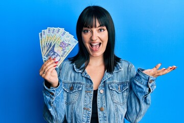 Young hispanic woman holding 100 romanian leu banknotes celebrating achievement with happy smile and winner expression with raised hand - obrazy, fototapety, plakaty