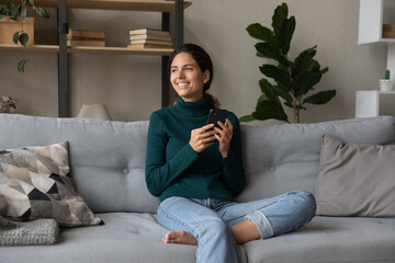 Leisure with smartphone. Happy confident young latin female sit on comfy sofa look to aside in dreamy mood distracted from cell device screen. Pleasant young female smiling browsing internet on phone