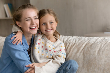 Happy dreamy loving young mother cuddling little cute child daughter, looking together in distance,...