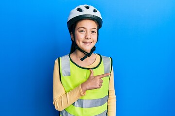 Beautiful brunette little girl wearing bike helmet and reflective vest cheerful with a smile of face pointing with hand and finger up to the side with happy and natural expression on face