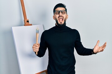 Handsome man with beard holding brushes close to easel stand crazy and mad shouting and yelling with aggressive expression and arms raised. frustration concept.