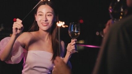 Young asian woman with sparklers is dancing and celebrating a new year. Fireworks, bengal lights in slow motion. Having fun at rooftop in the city.Happy interracial couple with glasses of wine .