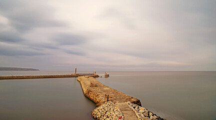 Two piers with lighthouses form a harbour entrance and the open sea is beyond. A headland is in the distance and a cloud filled sky is above.