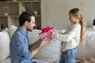Joyful cute small child daughter giving wrapped gift box to surprised young father, congratulating with happy birthday or International Father's Day, celebrating special event together at home.