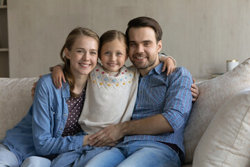 Portrait of happy bonding young couple parents cuddling little adorable kid daughter, relaxing on comfortable couch, enjoying carefree pastime together at home, loving family relations concept.