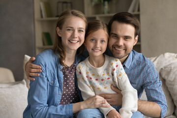 Happy bonding affectionate young couple parents cuddling little smiling adorable kid daughter sitting on comfortable couch, looking at camera, enjoying leisure weekend time together at own home.