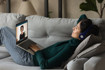 Just us girls. Millennial latin woman lie on couch in headset hold laptop on knees chat talk gossip...