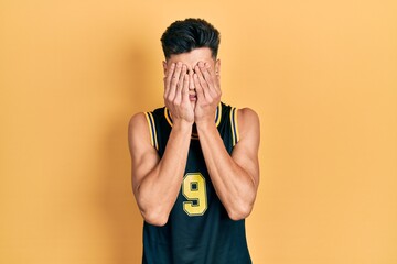 Young hispanic man wearing basketball uniform with sad expression covering face with hands while...