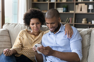 Happy mixed race guy using smartphone, confused suspicious girlfriend looking at boyfriend phone screen. Couple using online app on cellphone, chatting on internet, shopping, making virtual order