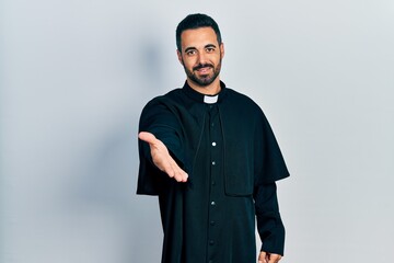 Handsome hispanic man with beard wearing catholic priest robe smiling cheerful offering palm hand...