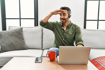 Young hispanic man with beard wearing call center agent headset working from home very happy and smiling looking far away with hand over head. searching concept.