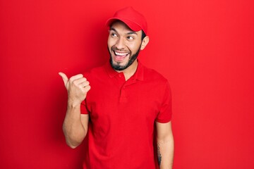 Hispanic man with beard wearing delivery uniform and cap smiling with happy face looking and pointing to the side with thumb up.